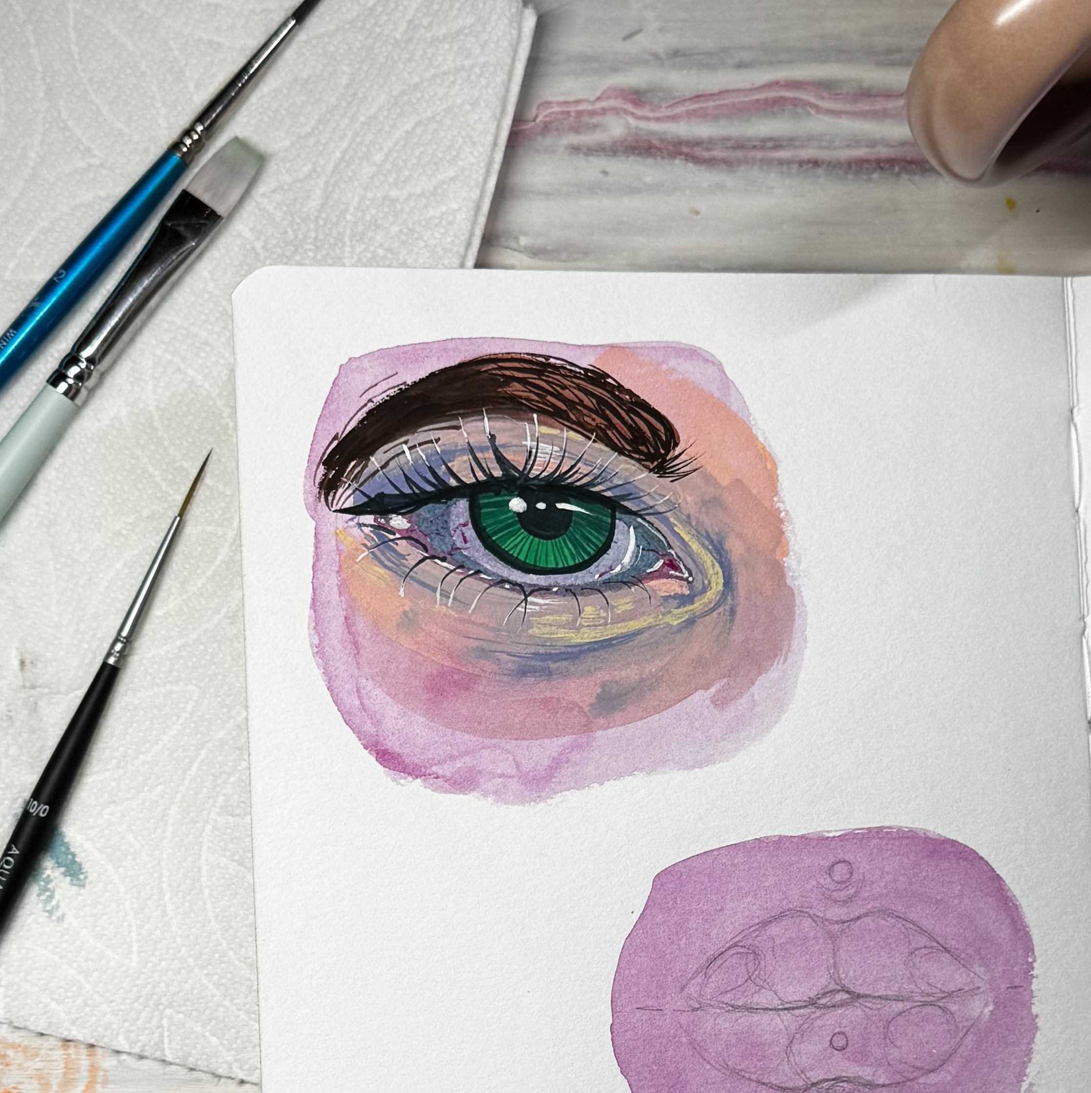 Artistic drawing of an eye with watercolor techniques on a piece of paper, accompanied by painting tools and a cup of tea.
